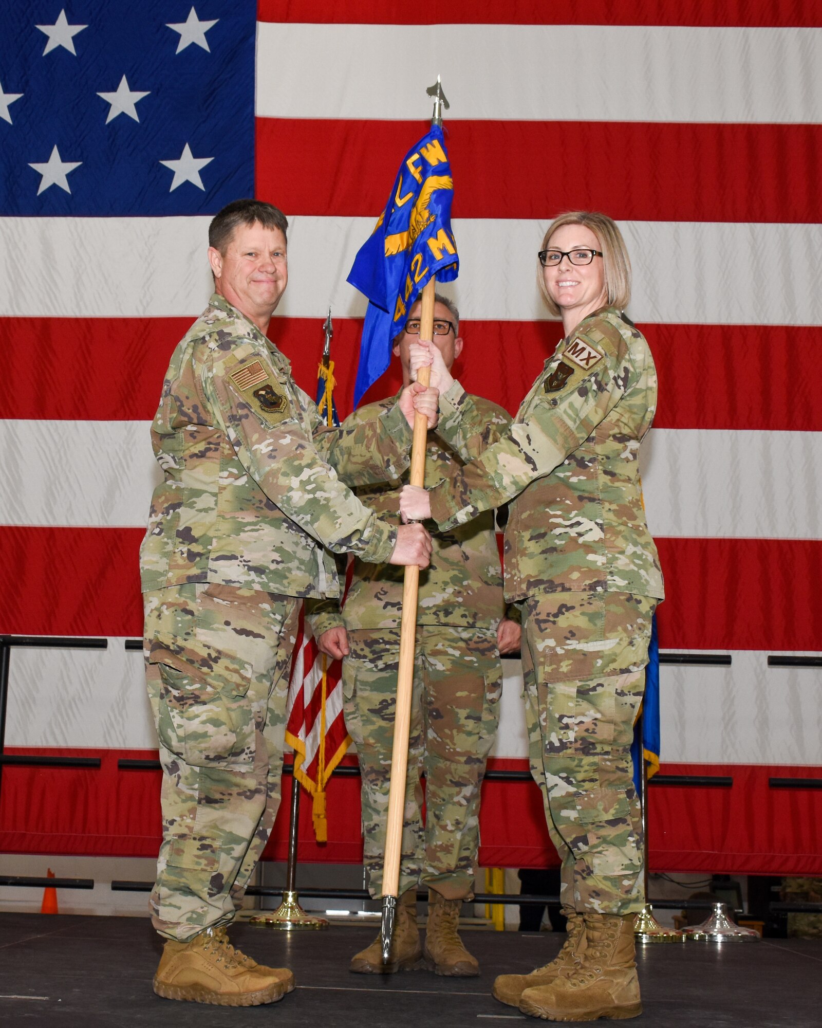 A man on the right hands a blue flag to a woman on the left. Both are wearing operational camouflage pattern fatigues and standing in front of a very large American flag.