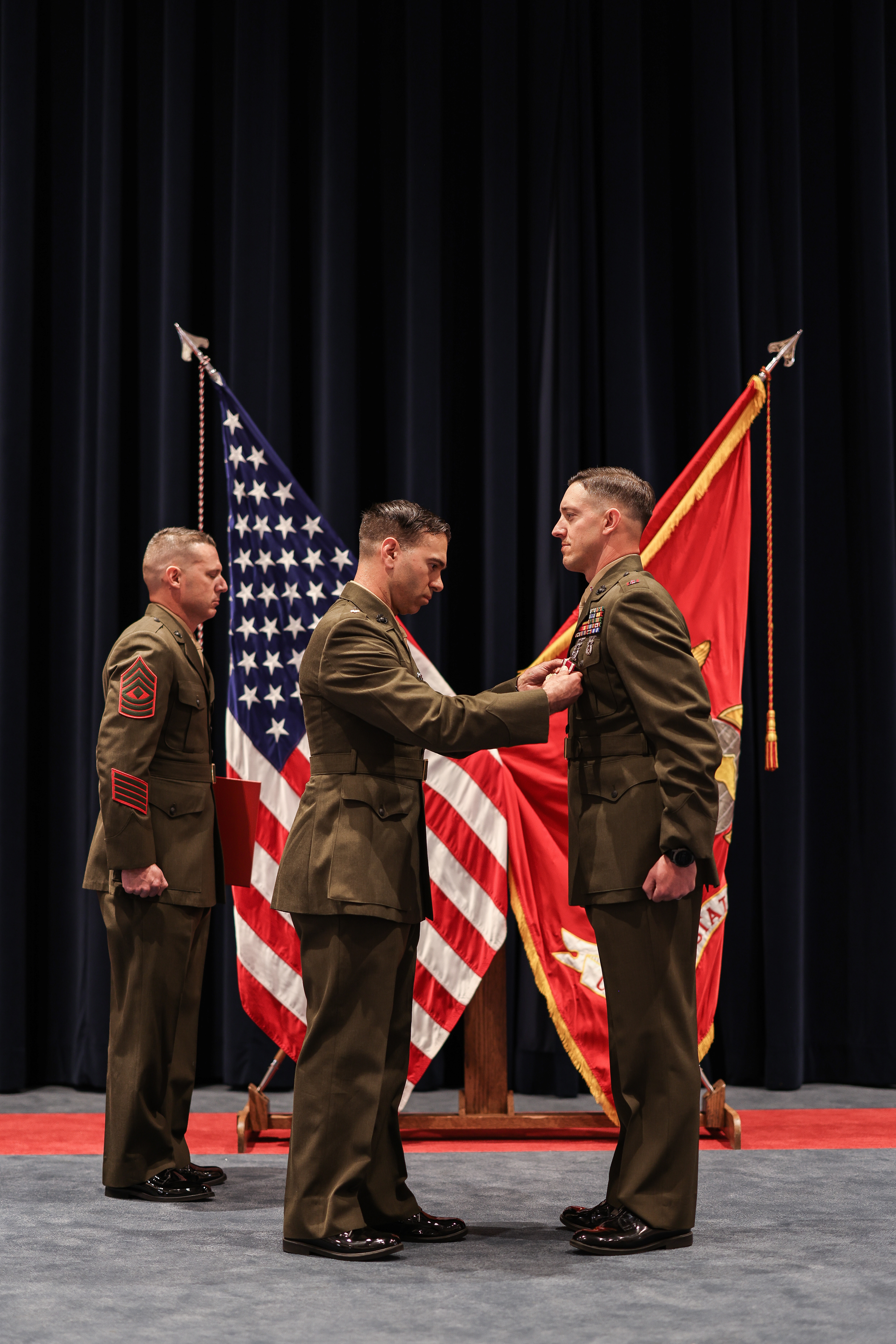 Three Marines on stage, one is pinning something on another's jacket.