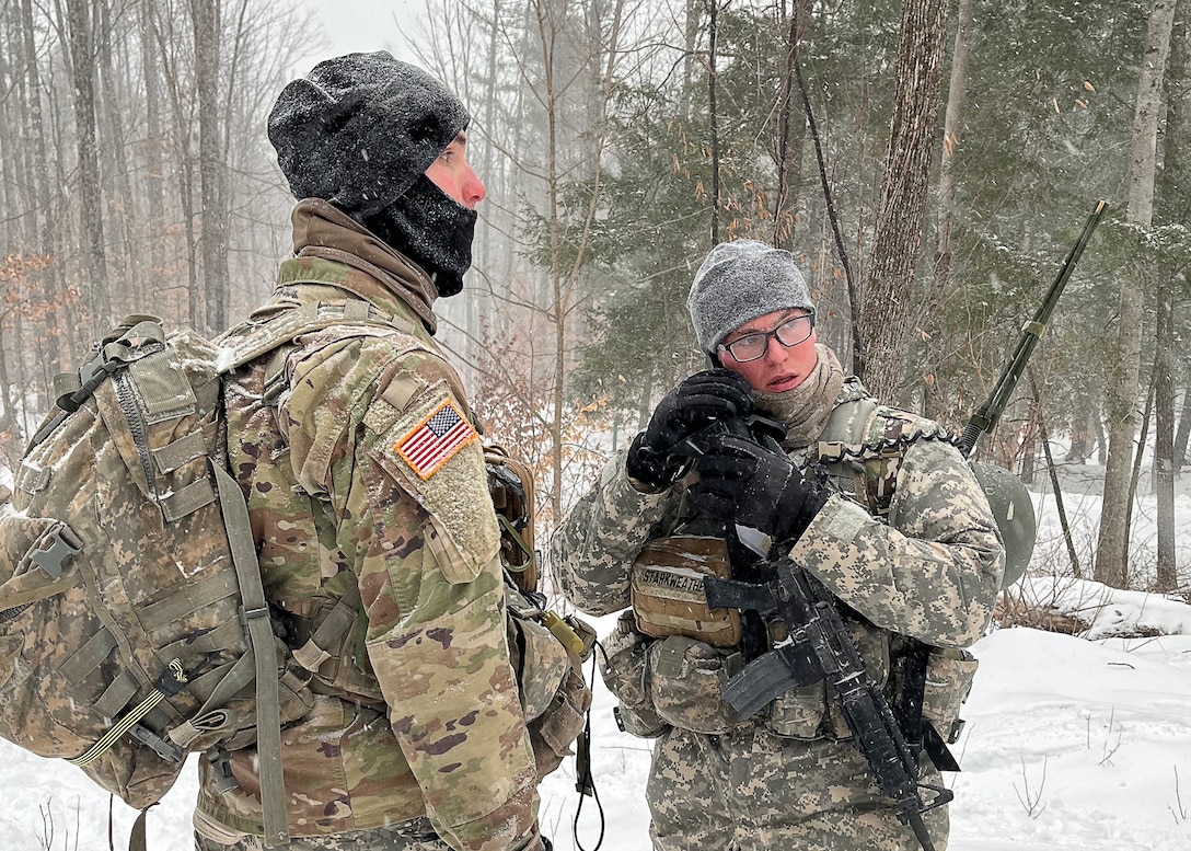 Two ROTC students dressed in Army winter uniforms, one of the students is using a radio. Snow is on the ground.