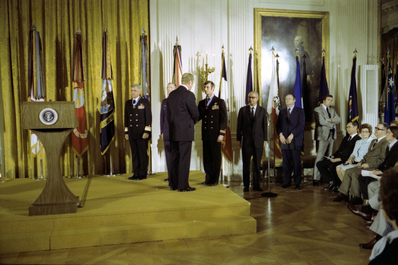 A man shakes hands with another man on a podium. A few others stand around them, as a few people sit in a crowd.