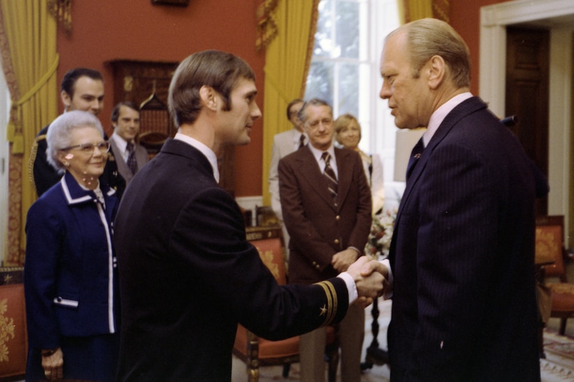 A man shakes hands with another man as a few others stand around them.