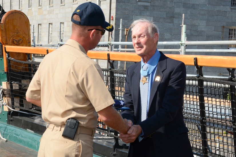 A man wearing a Medal of Honor shakes hands with a sailor.