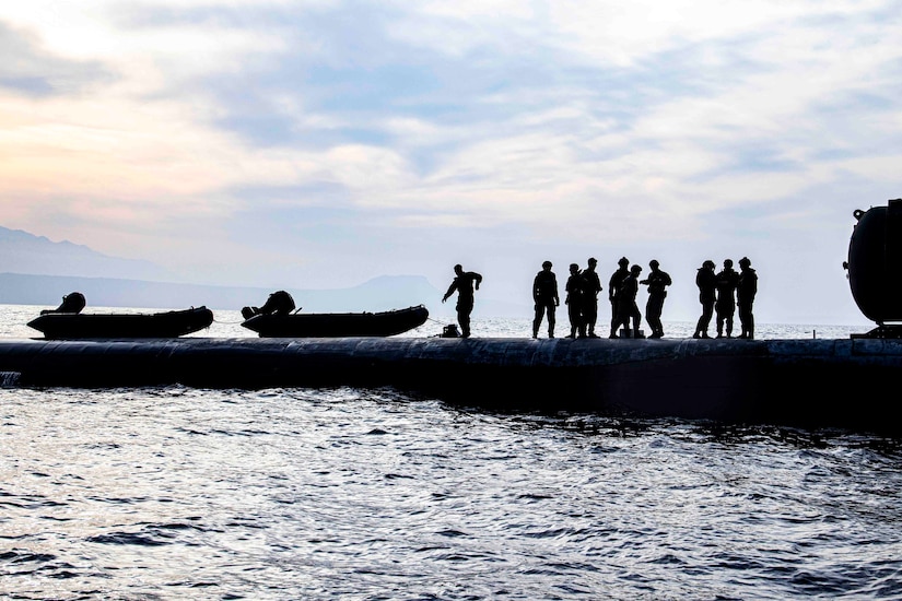 Marines stand on top of a submarine next to two small inflatable boats.