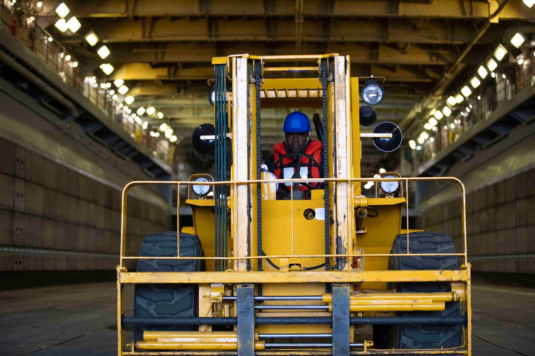 A sailor drives a forklift in the well deck of a ship.