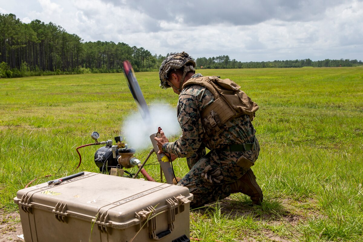 A servicemember kneels in a grassy area and operates military equipment.