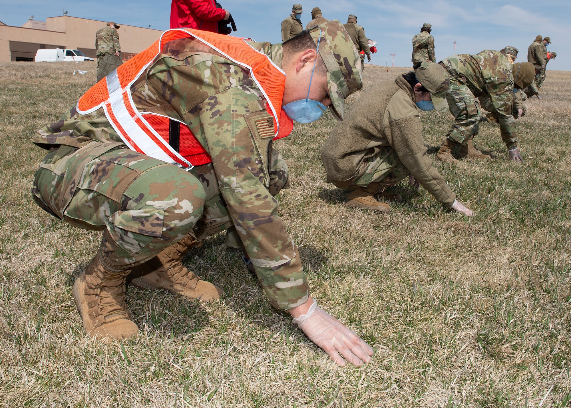 a diagonal line of Airmen in uniform crouching down as they search grassy area in front of them
