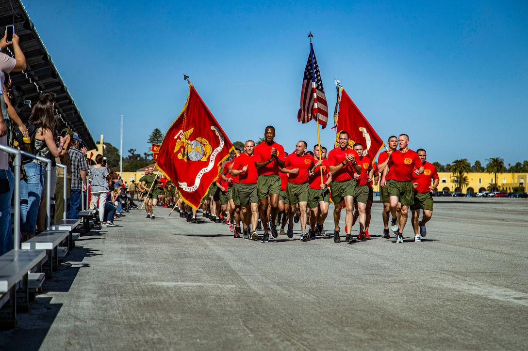A group of Marines run in formation while an audience watches.