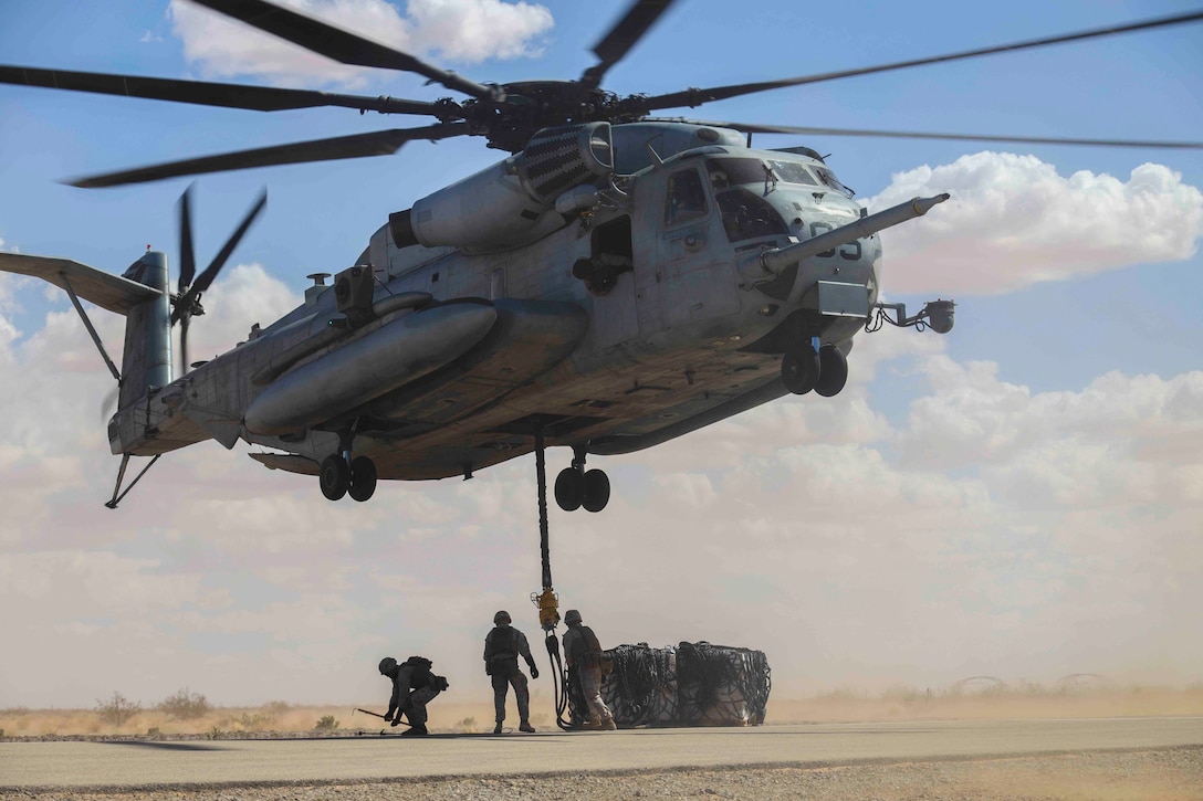 Marines prepare to attach cargo to an airborne helicopter.