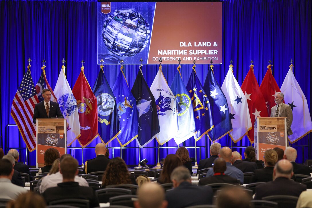 Two people at lecterns on stage with flags in the background.