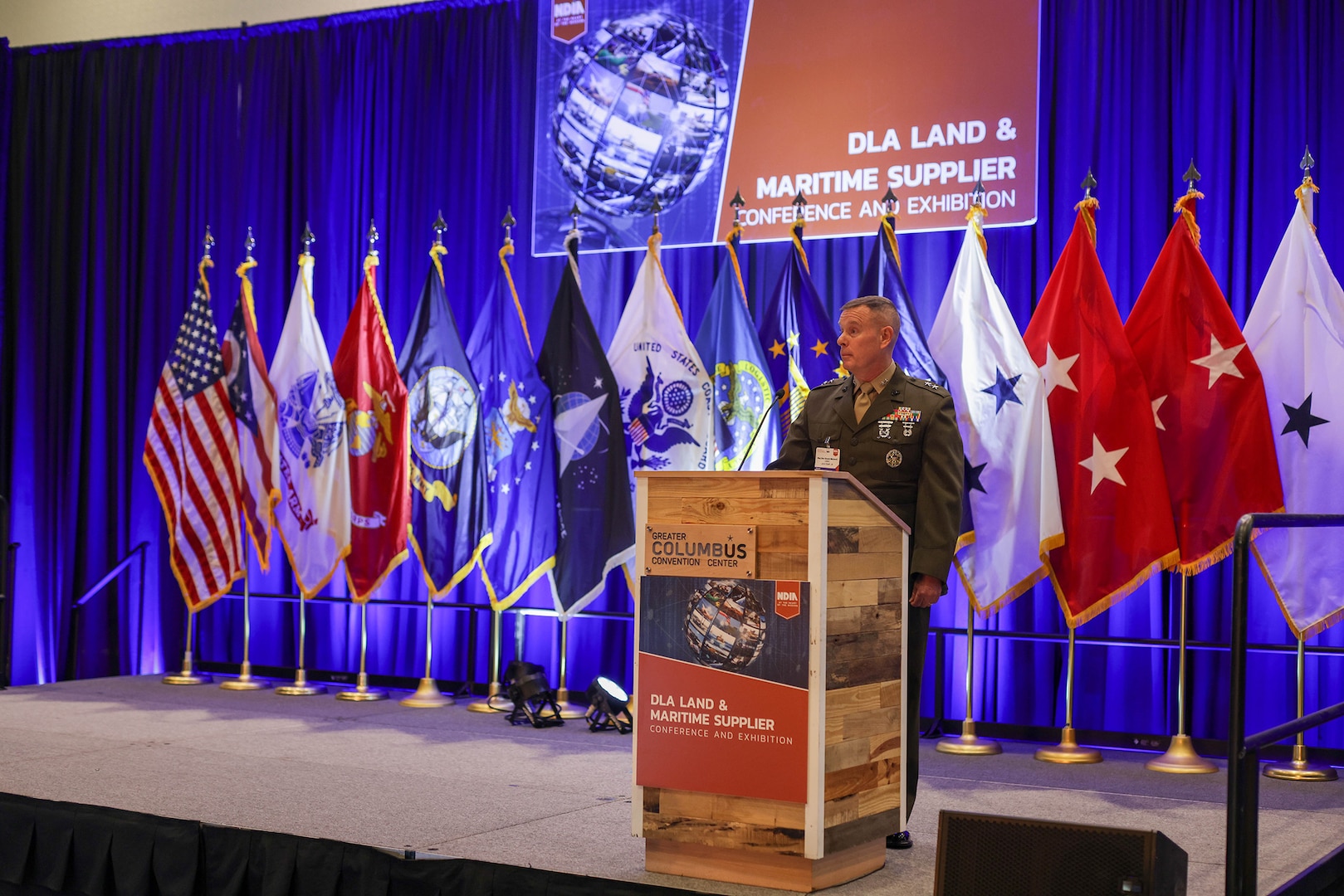 Man in US Marines dress uniform on stage with flags in the background.