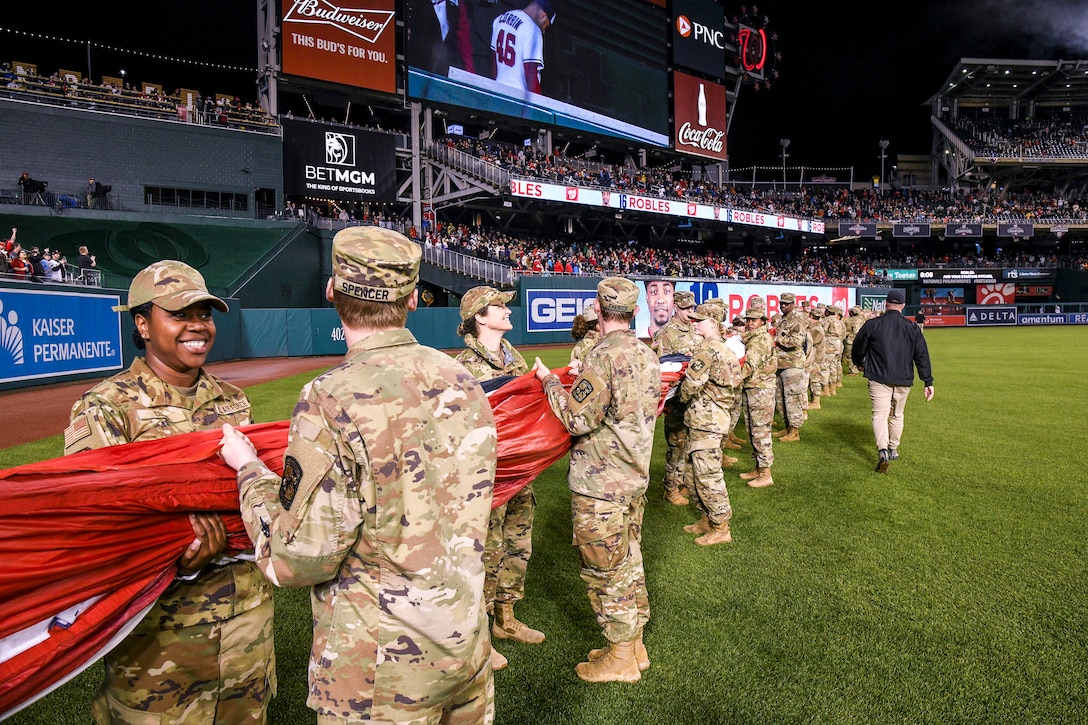 Airmen and soldiers stand in a long row on a ballfield holding a furled American flag.