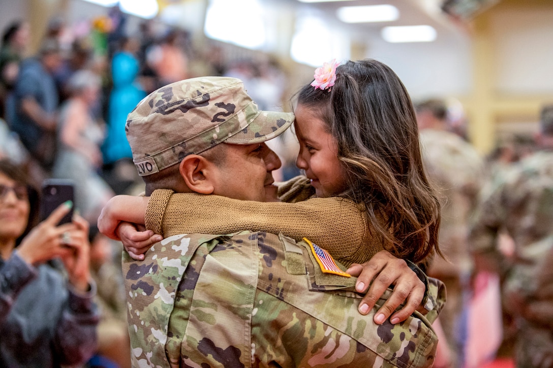 A smiling soldier holds a little girl as the two look at each other.