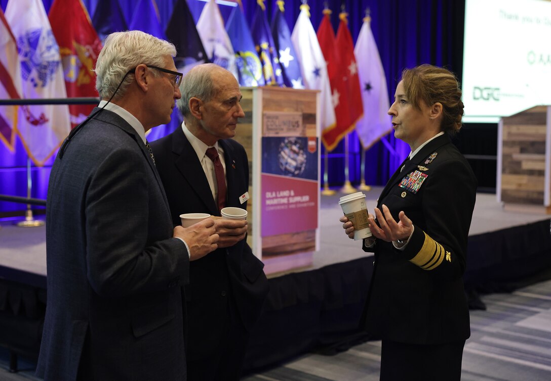 Woman in military uniform speaks to two men in business suits.