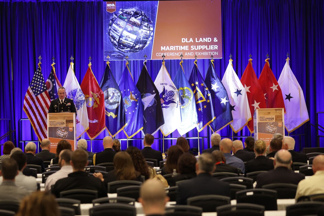 Man in uniform speaks at a podium on stage with flags behind him.