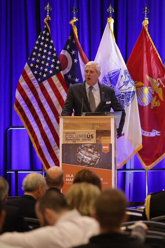 Man in business suit speaks at a podium on stage with flags in the background.