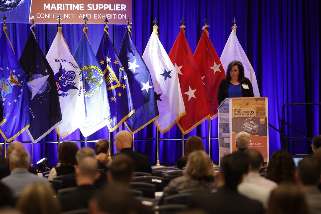 Woman in a business suit on stage at a podium with flags behind her.