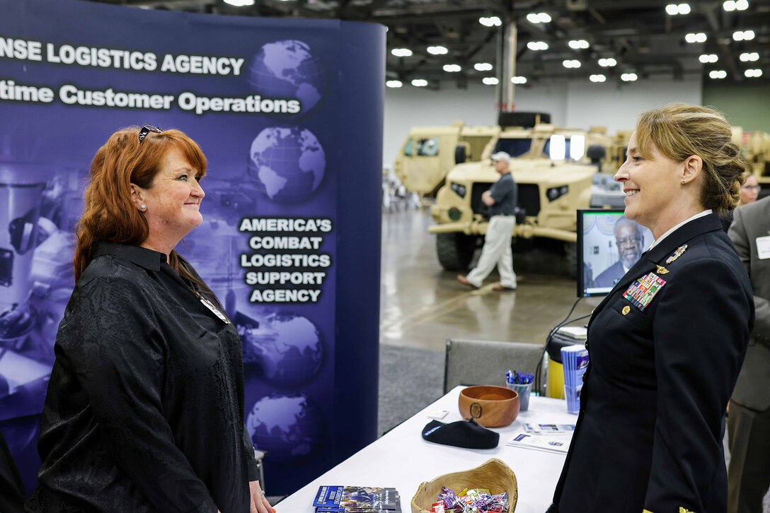 Woman in military uniform greets another woman at a booth at a exhibition.