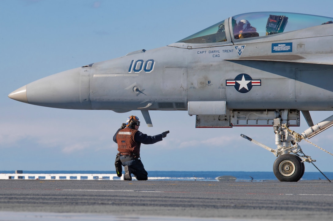 A sailor kneels while signaling an aircraft on the deck of a ship.