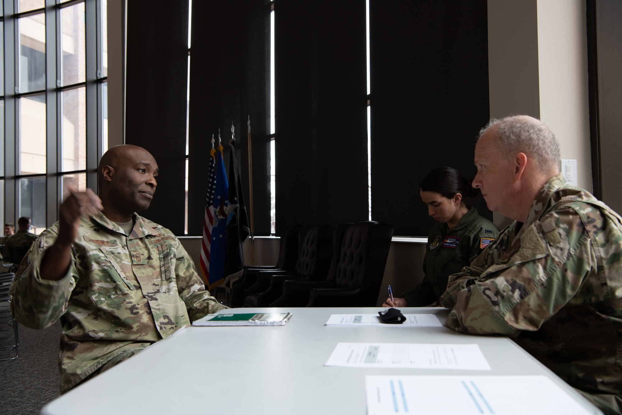 Two men sit at a table across from each other having a conversation while a woman in aircrew uniform takes notes in the background.