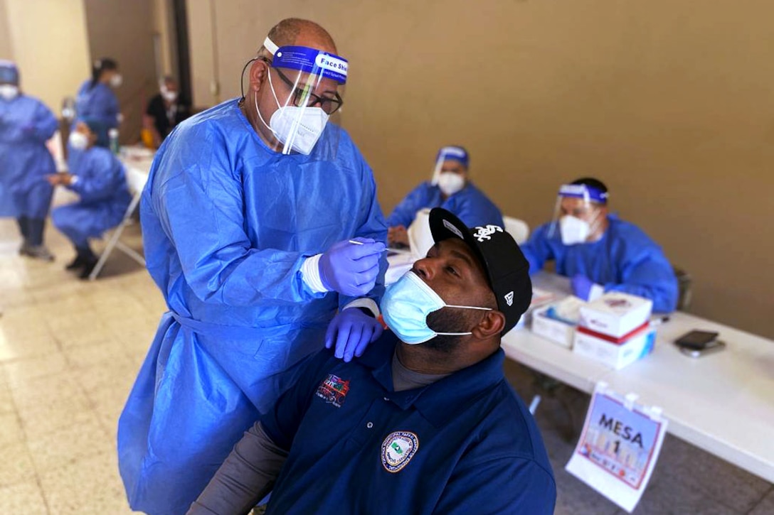 A man wearing personal protective equipment holds a nasal swab to the nose of a man seated in a chair.