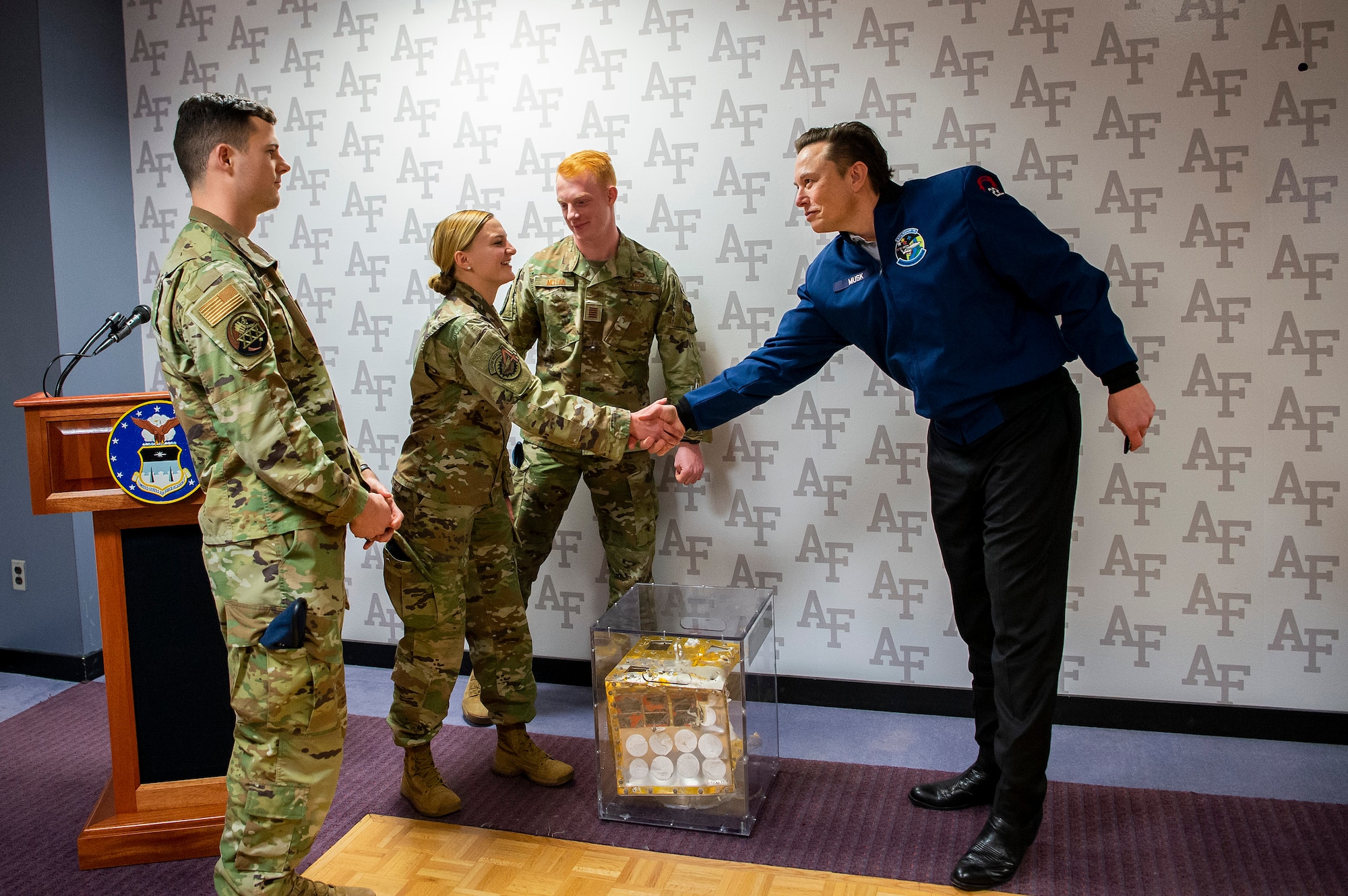 Elon Musk, Space X CEO, standing in a room with three Air Force Academy cadets who are presenting a capstone project to him.