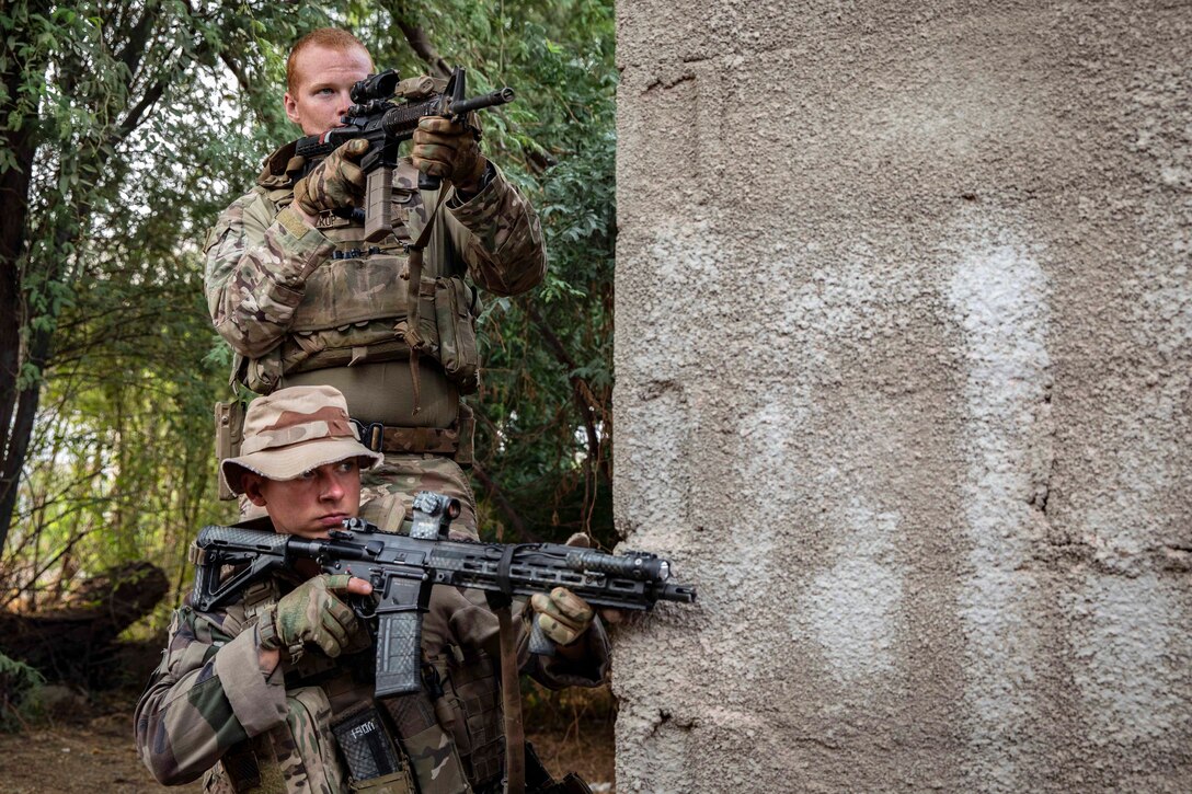 U.S. and French troops hold weapons while standing near a concrete structure in the woods.