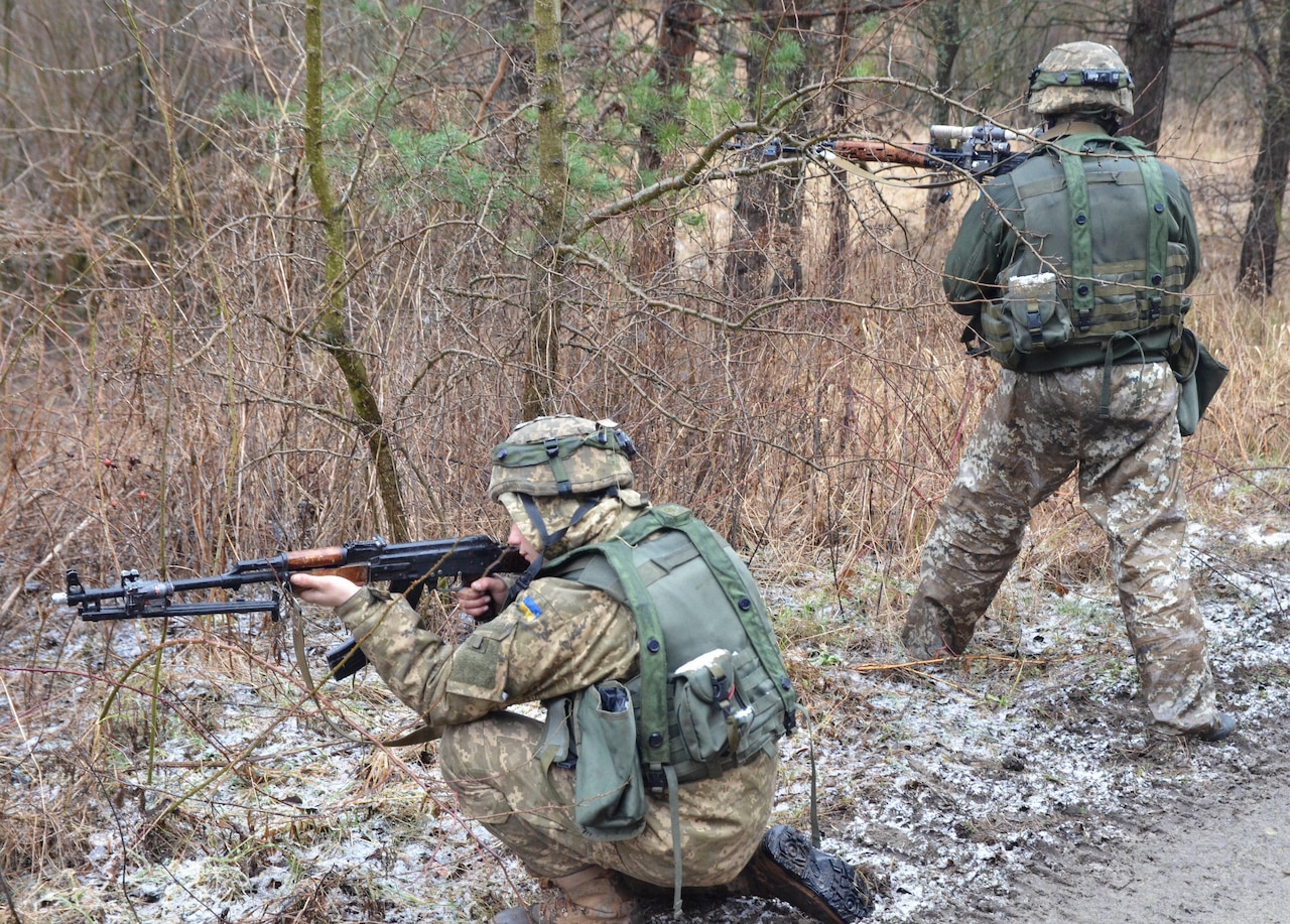 One soldier kneels outdoors and points a rifle, while another stands.