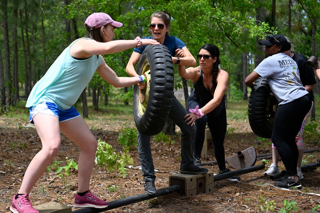 Civilians handle a tire while balancing on a raised bar in the woods.