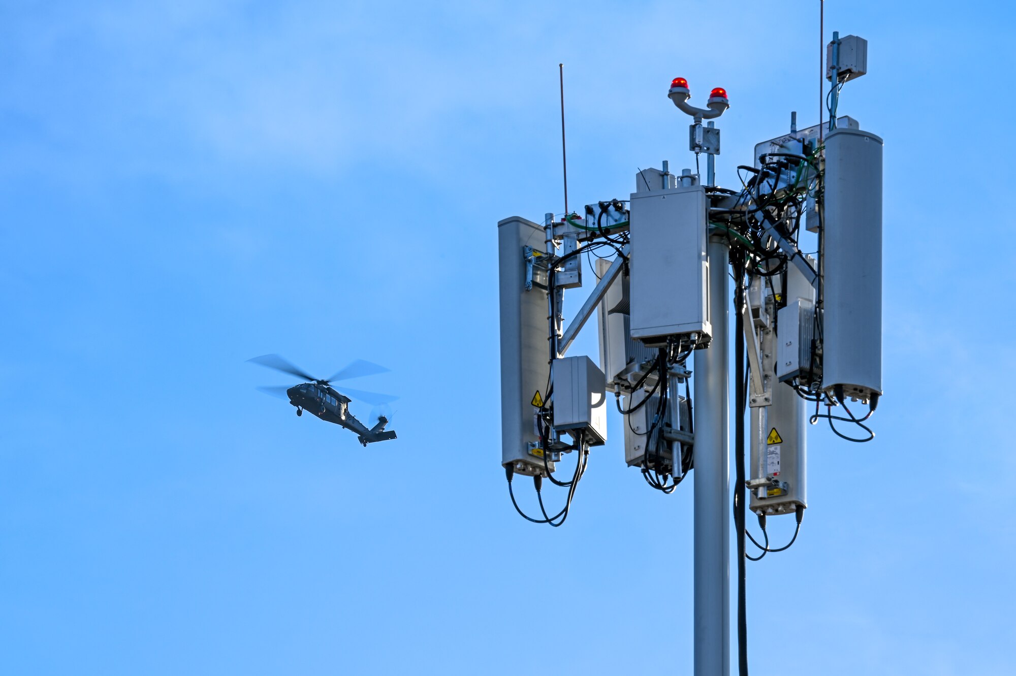 An UH-60 Blackhawk flies over the antennae of a mobile test station in blue sky conditions.