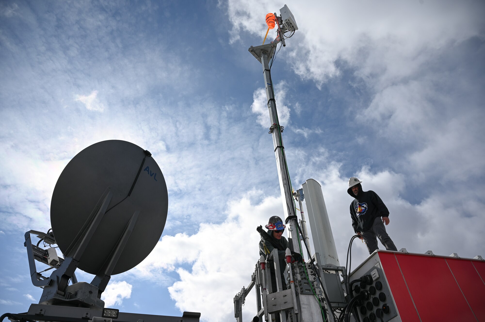 A work crew stands on top of a 5G mobile test station  with white cloud filled blue sky in the background.