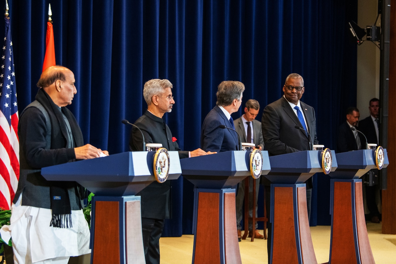 Four men stand on a stage with flags behind them.