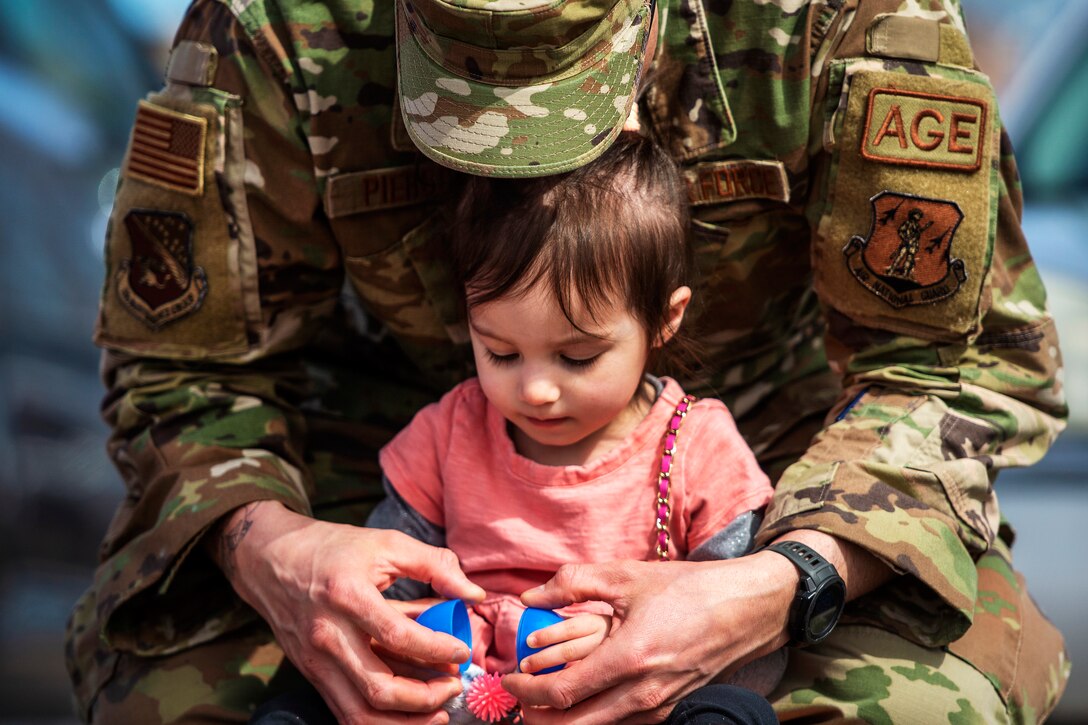 An airman cradles a child from behind while opening an Easter egg.