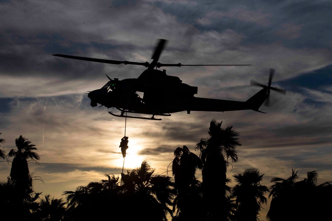 A Marine shown in silhouette fast-ropes out of an airborne helicopter.