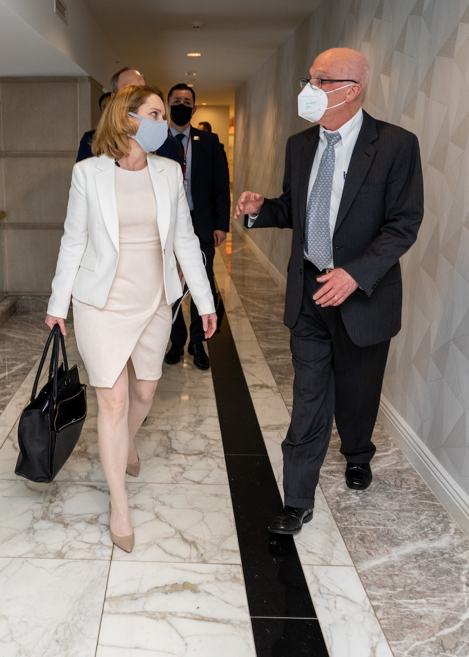 A woman and a man, each wearing face masks, walk down a hallway together.