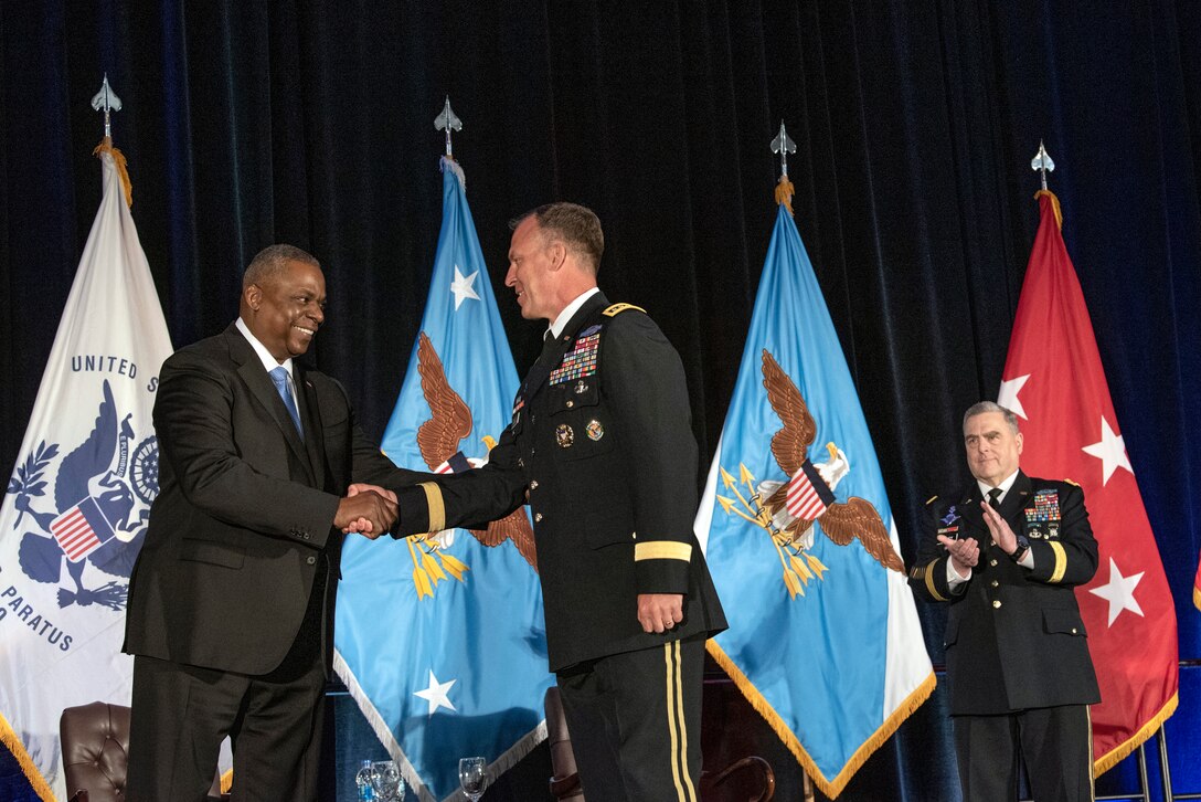 Secretary of Defense Lloyd J. Austin III shakes hands with a commander on a stage with the Chairman, with flags in the background.
