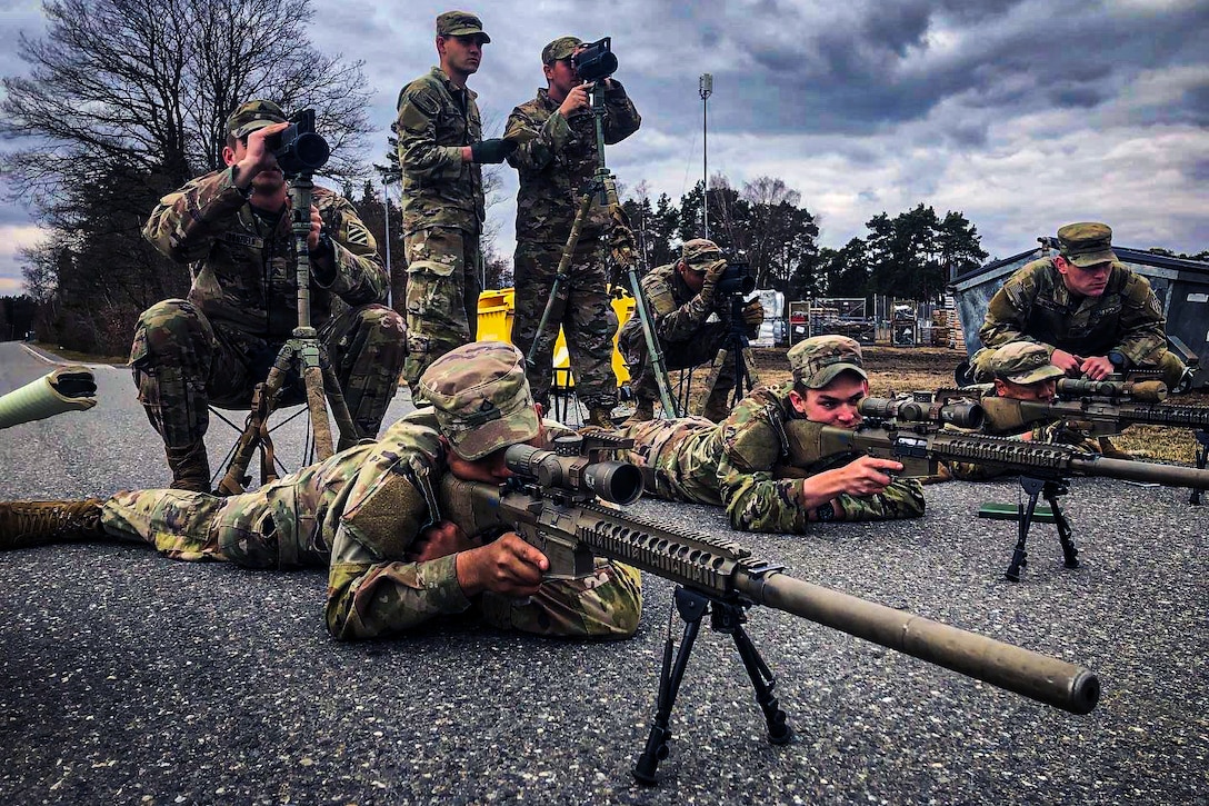Three soldiers lay on the ground holding rifles during sniper section conduct range estimation and target detection training while five other soldiers observe.