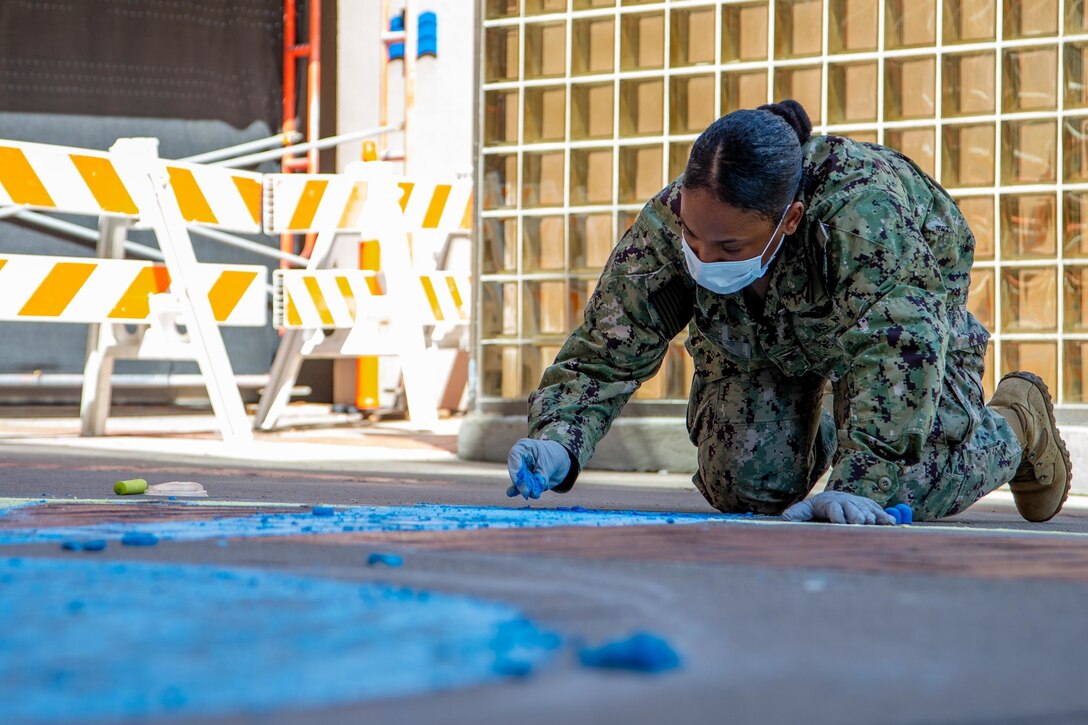 A sailor kneels on the ground and draws a picture with chalk.
