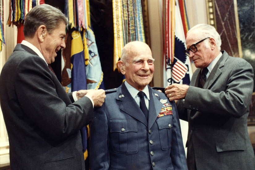 An older man in a military dress jacket smiles as two other men pin items to his epaulettes.