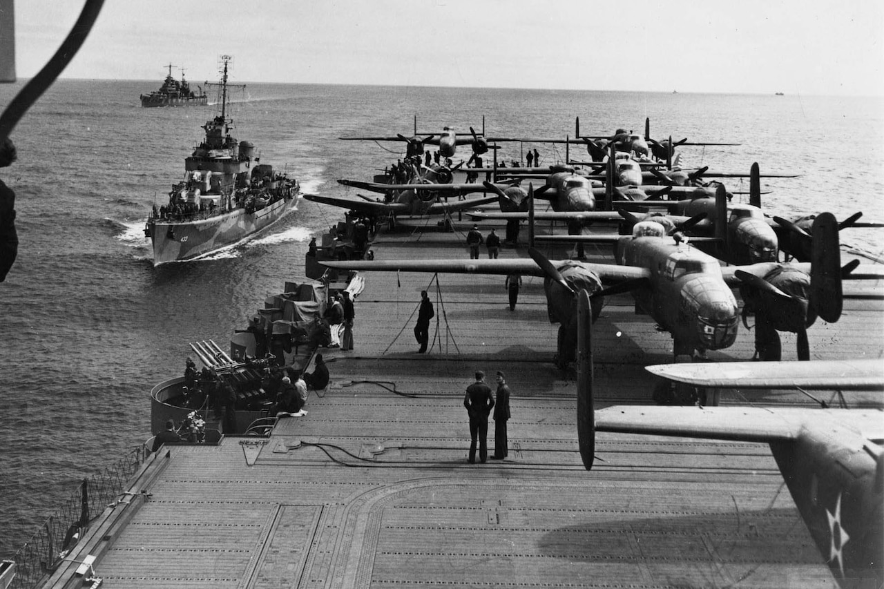 Several airplanes sit on the deck of a ship while two other ships follow in its wake.
