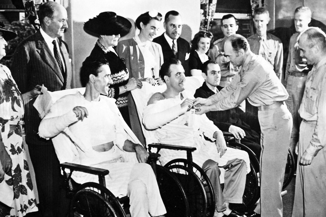 Two men in wheelchairs are presented with pins as several others stand behind them to watch.