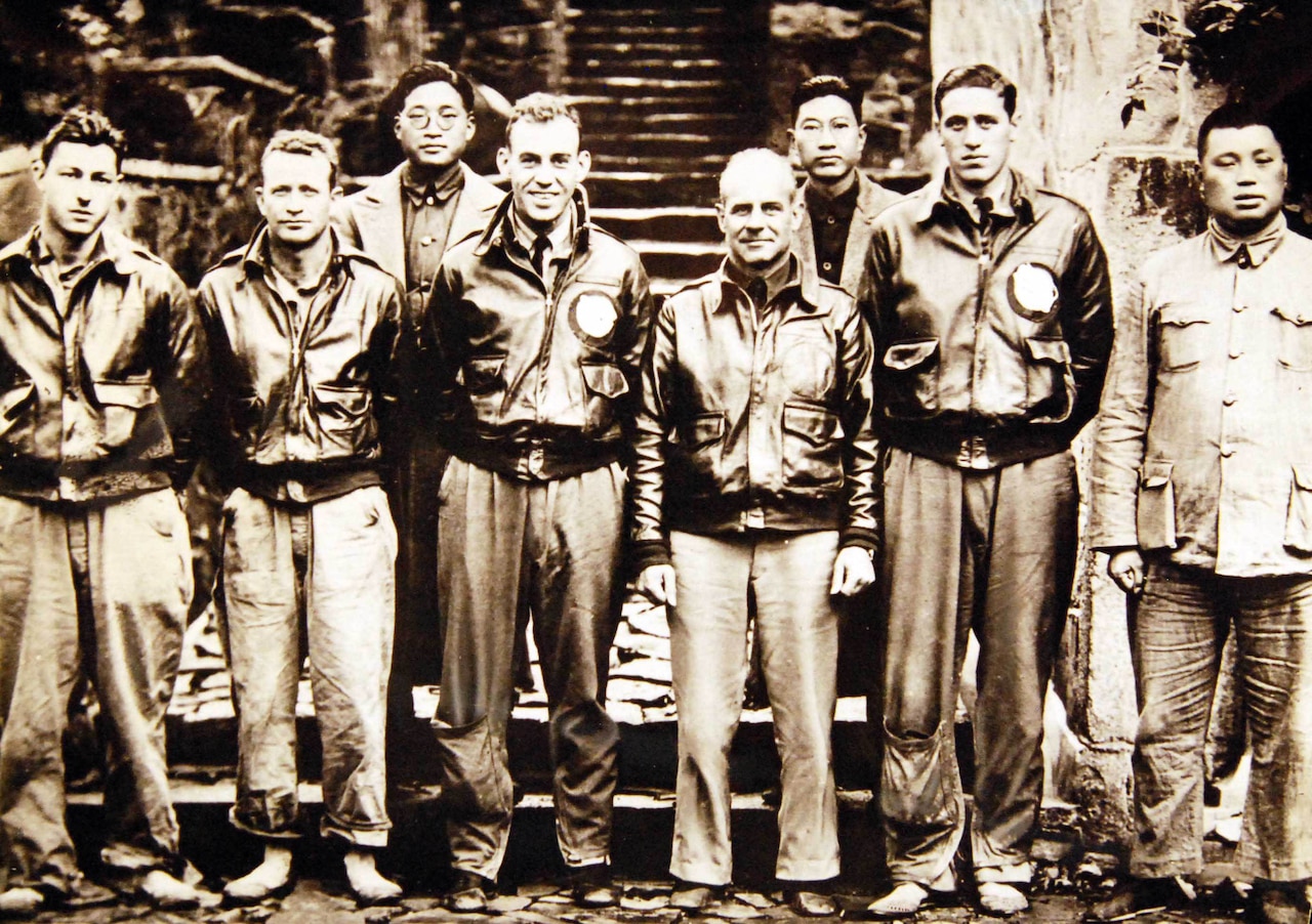 Eight men, five of whom are wearing bomber jackets, pose for a photo.