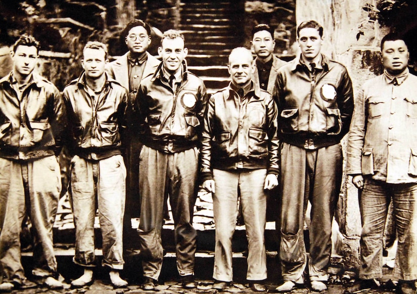 Eight men, five of whom are wearing bomber jackets, pose for a photo.