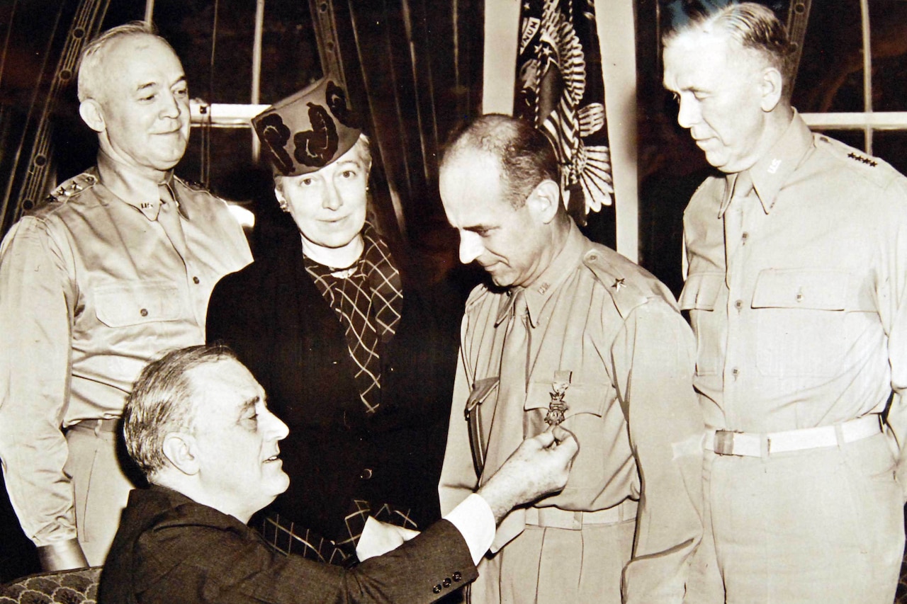 A seated man adjusts the pin on the lapel of a standing man’s jacket. Two men and a woman stand beside them.
