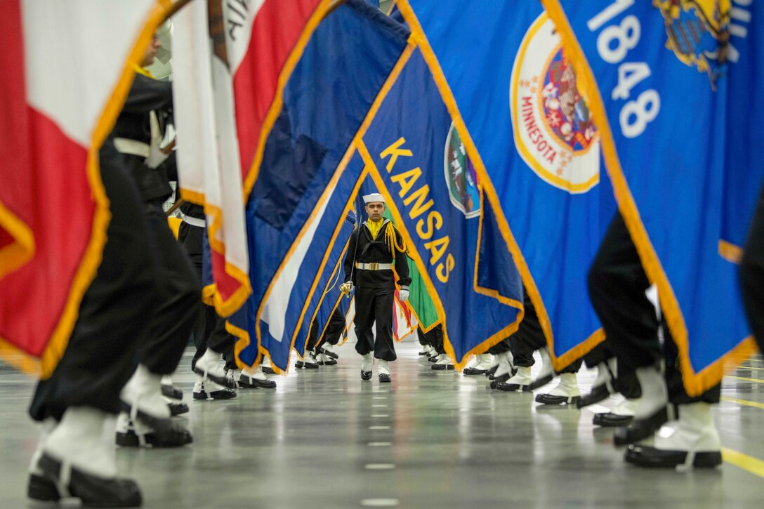 Navy recruits carrying flags march in a parallel line as a fellow recruit walks down the center.