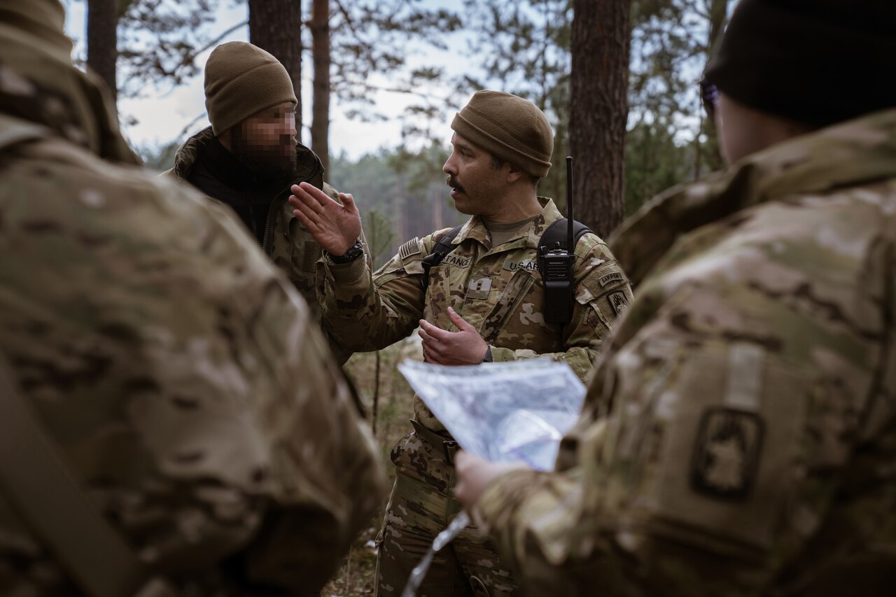 A Soldier talks to a digitally obscured person in a forest setting near more soldiers.