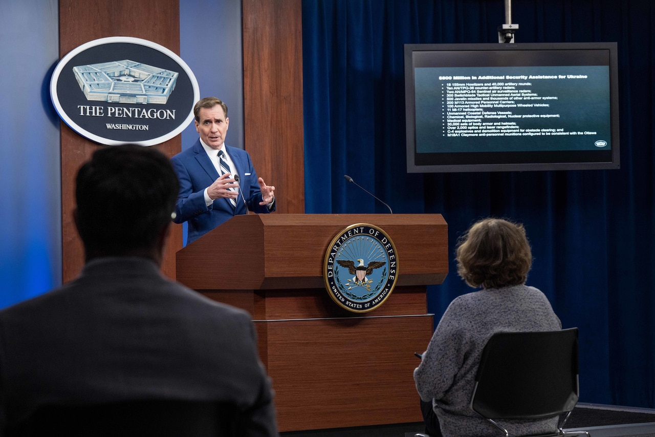 Pentagon Press Secretary John F. Kirby speaks to a group of people from a lectern with a tv monitor showing info on the side.