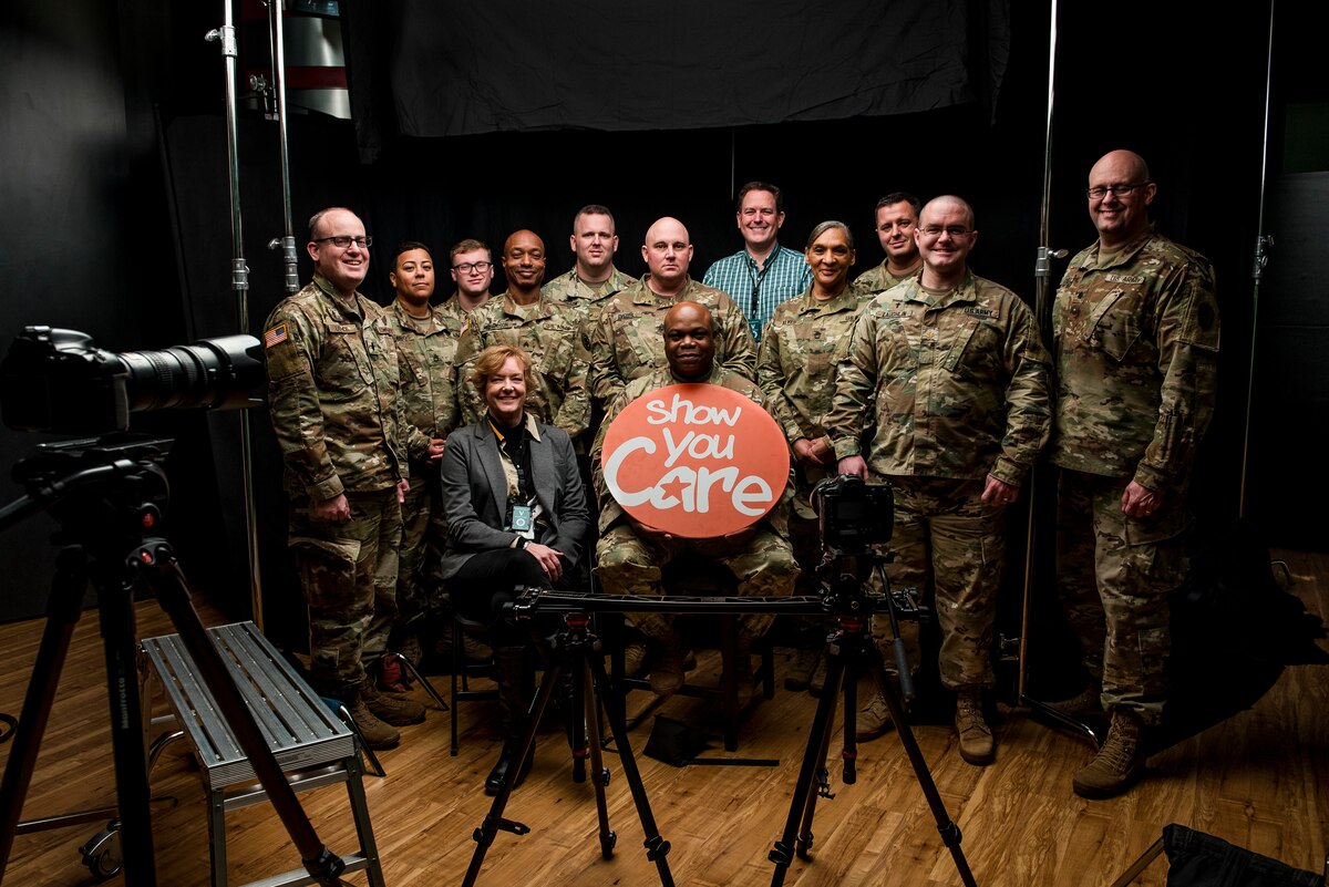 Soldiers pose for a group photo while a man at center holds a sign that reads: Show you care.