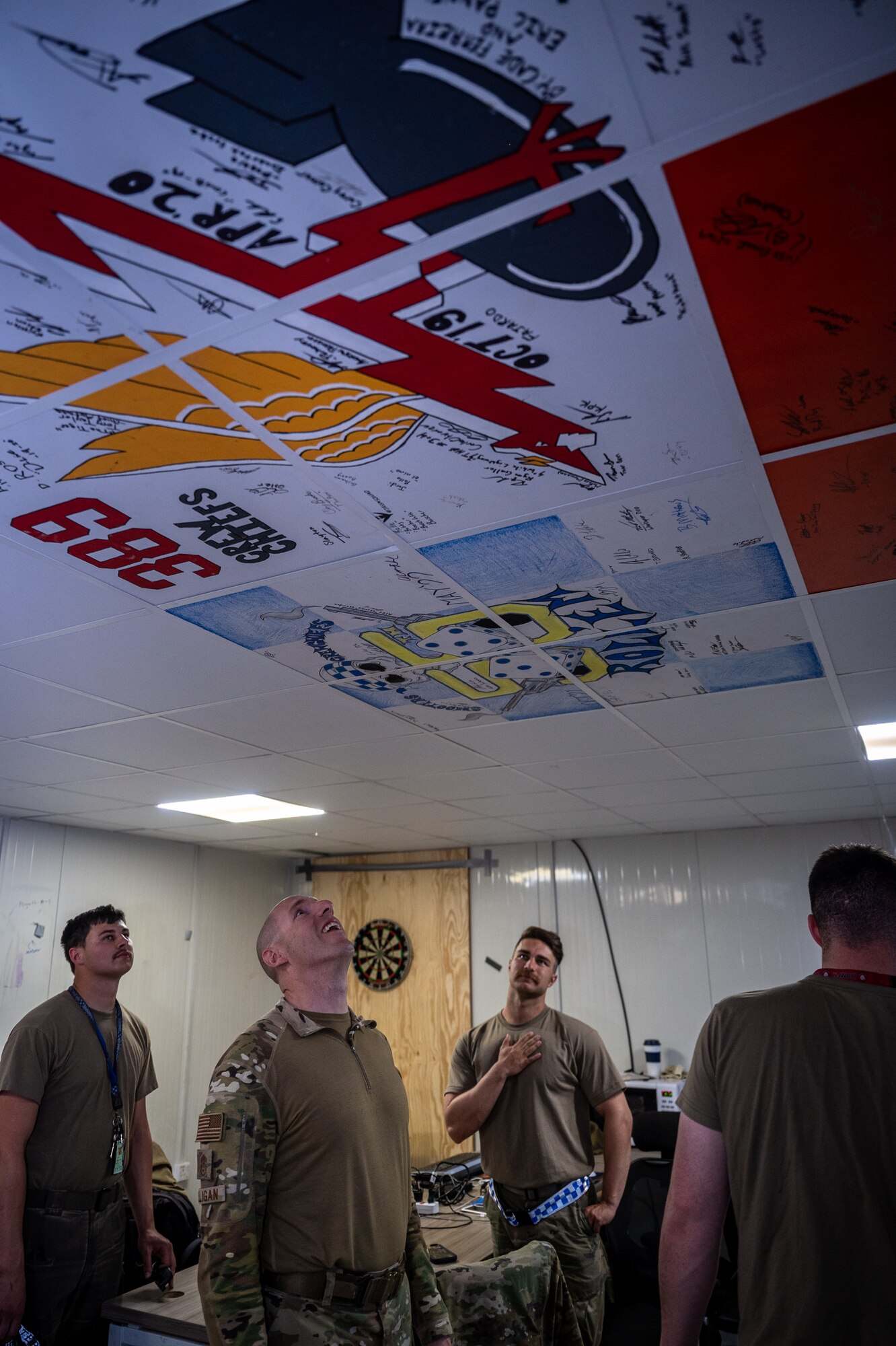 Team members from the 55th Expeditionary Fighter Generation Squadron point out several ceiling murals to Chief Master Sgt. Sean Milligan, 332nd Air Expeditionary Wing command chief, inside the crew chief lounge and office area at an undisclosed location in Southwest Asia, April 9, 2022. The heritage ceiling tiles were decorated by EFGS crew chiefs. (U.S. Air Force photo by Master Sgt. Christopher Parr)