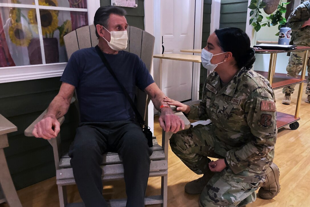 An airman wearing a face mask kneels while speaking to a man wearing a face mask sitting in a chair.
