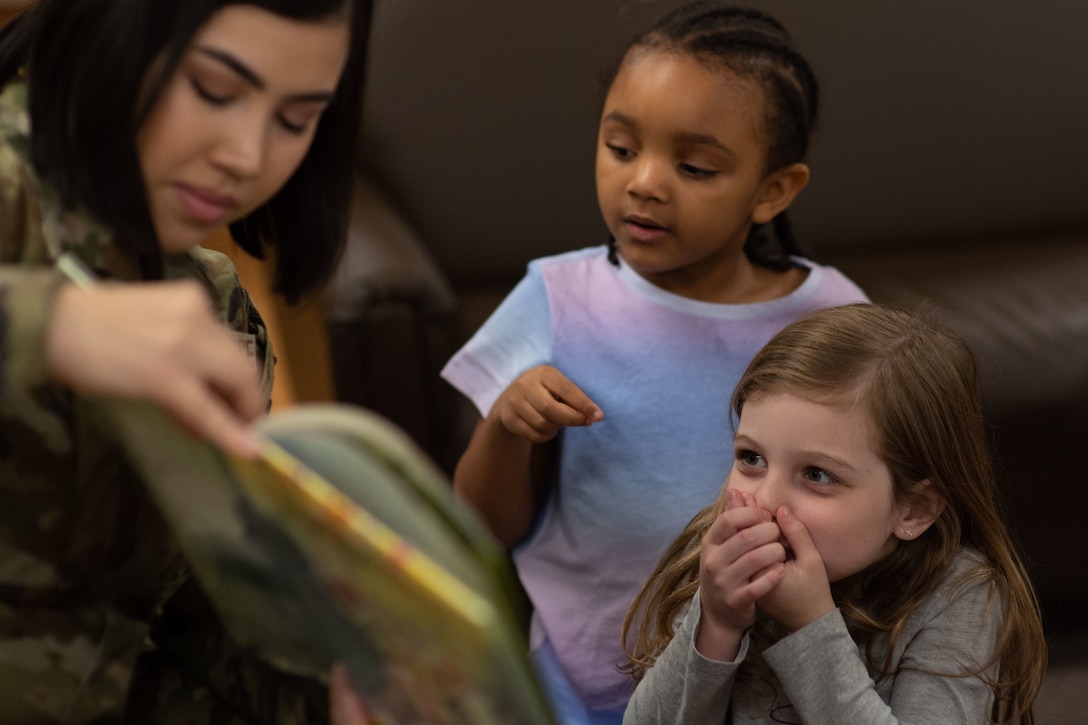 Two children listen and react as an airman reads them a story.